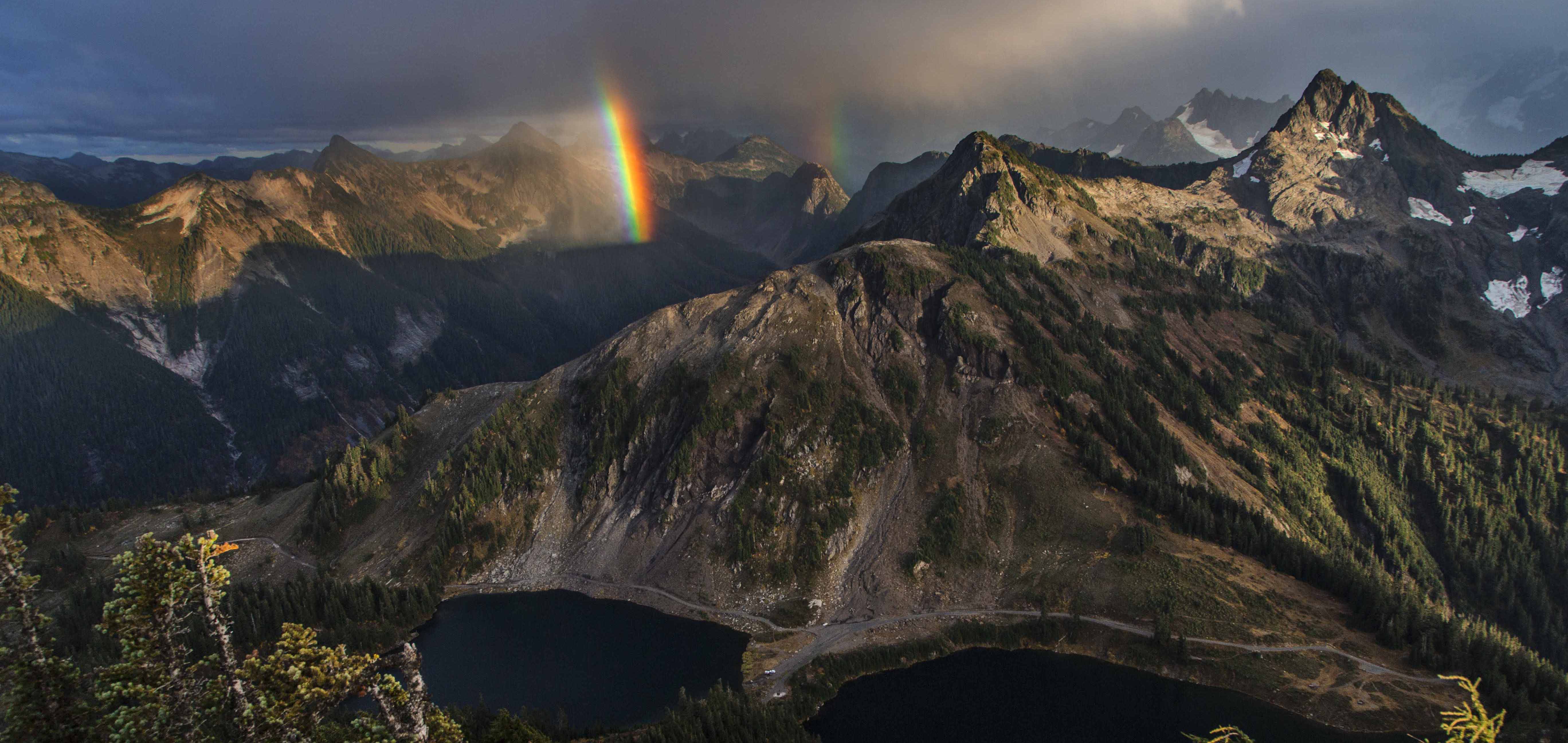 A lake rainbow disappears into jagged peaks, capped with snow. Below the peaks is a dark lake. The lake is bordered by a thin ribbon of a road.