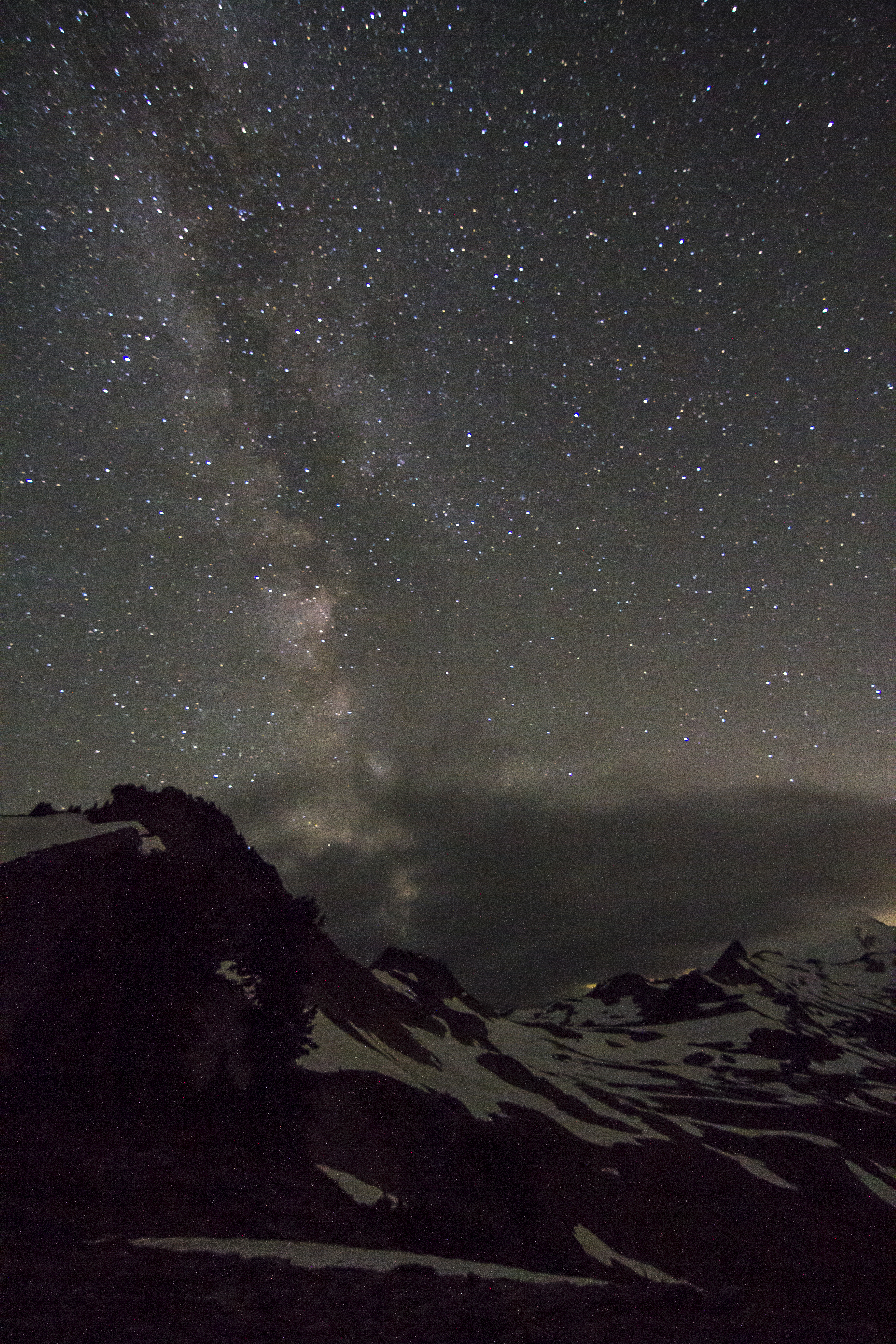 A panoramic shot of a snow covered mountain beneath the night sky. The sky is illuminated with thousands of stars, and you can see an arm of the Milky Way